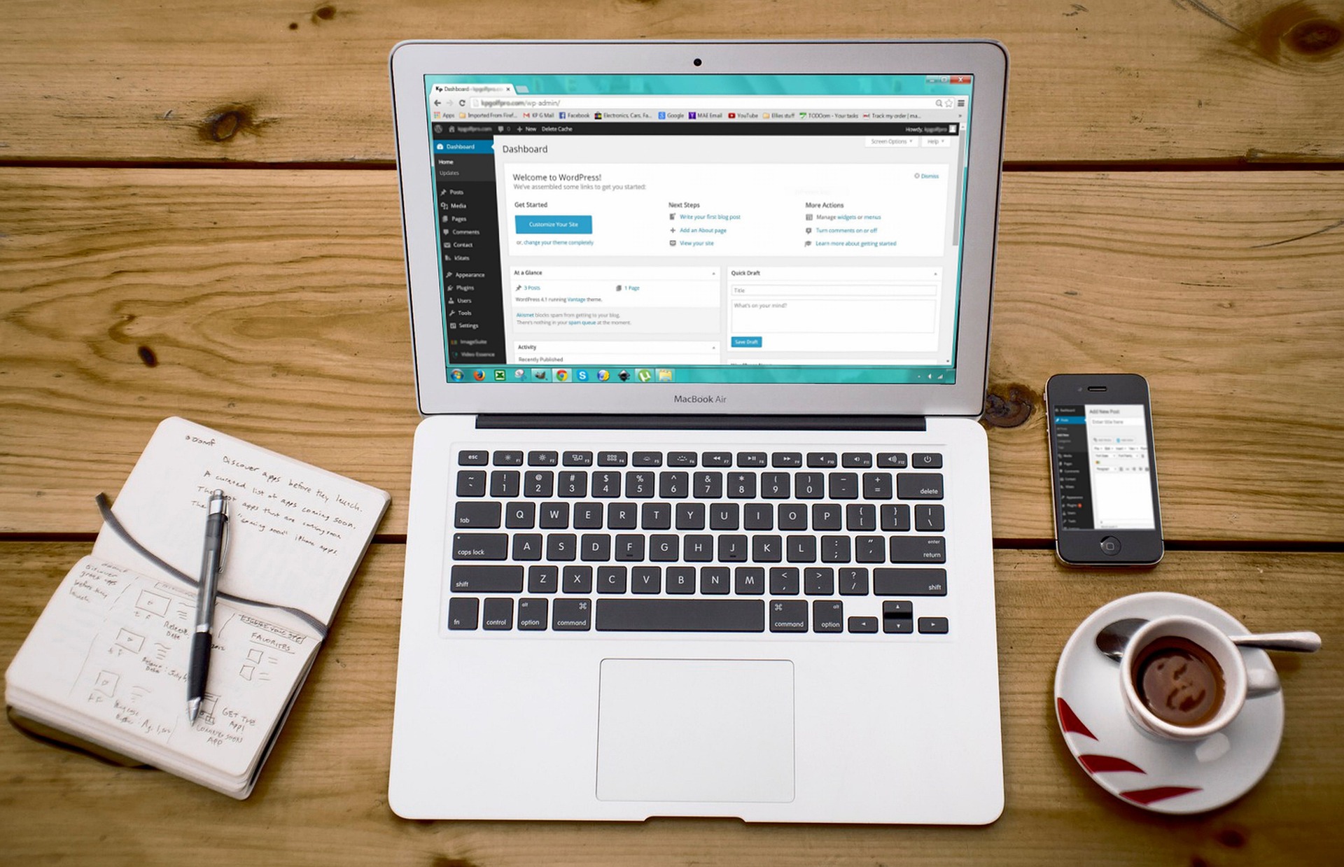 laptop sitting on table with notepad and pen on the left and a cup of espresso and a cell phone on the right